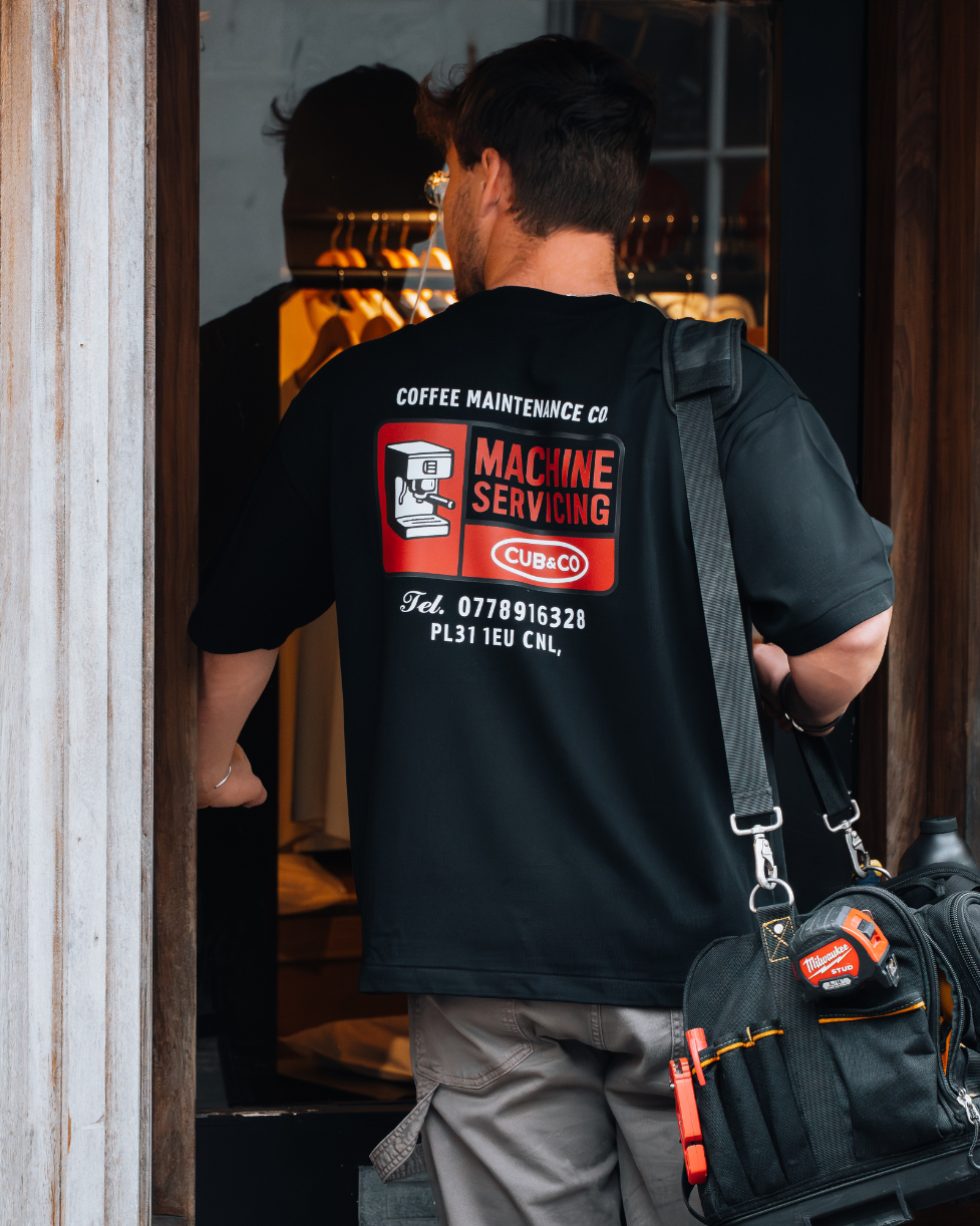 Person wearing a black t-shirt with a red and white graphic on the back, carrying a tool bag.