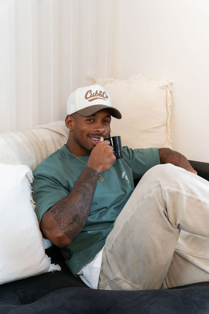 Man sitting on a couch wearing a cap and green shirt, holding a cup.
